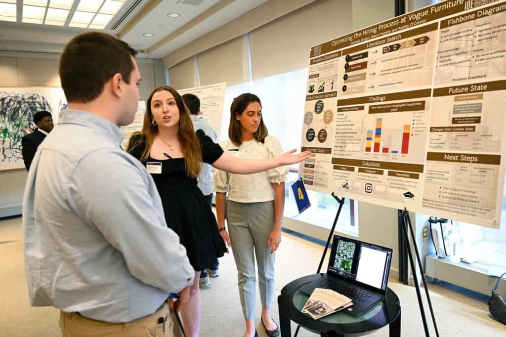 A student gestures at a poster and speaks to other students in a senior design presentation.