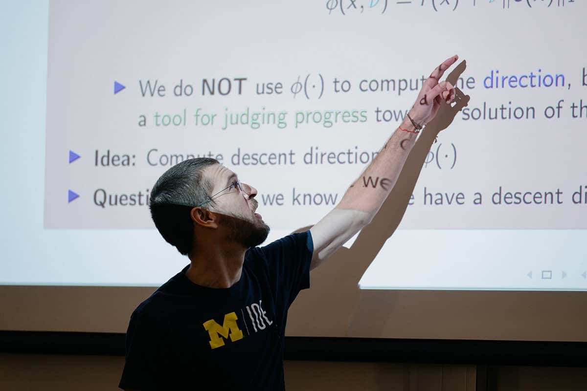 A person wearing a U-M IOE tshirt points at an equation on o projector screen in a dark room.