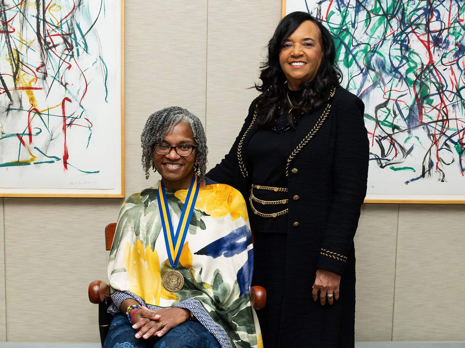 One woman sits in a chair with a medal one while the other woman smiles standing next to her