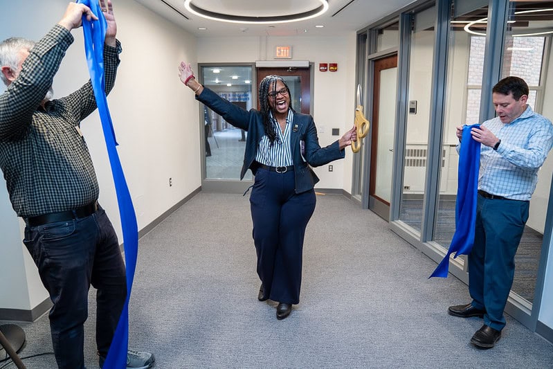 Julie Simmons Ivy cheers with hands in the air as she holds oversized scissors. Two people to either side collect the ribbon she just cut.