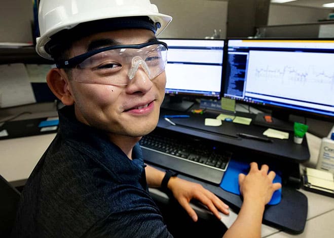 Person in safety glasses and hard hat on a computer in a cubical